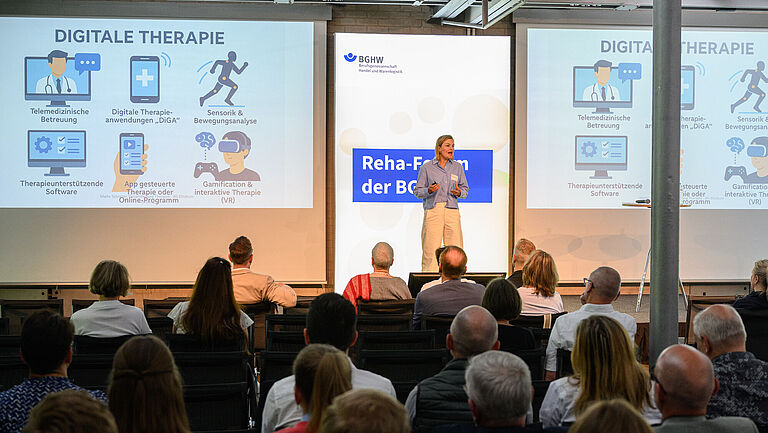 Blick ins Plenum des Reha-Forums: Eine junge Frau steht gestikulierend auf der Bühne, mit dem Gesicht zur Kamera. In der Präsentation hinter ihr kann man "Digitale Rehabilitation" lesen. Im Vordergrund sind die Rücken der Zuschauer zu sehen, die vor ihr sitzen.
