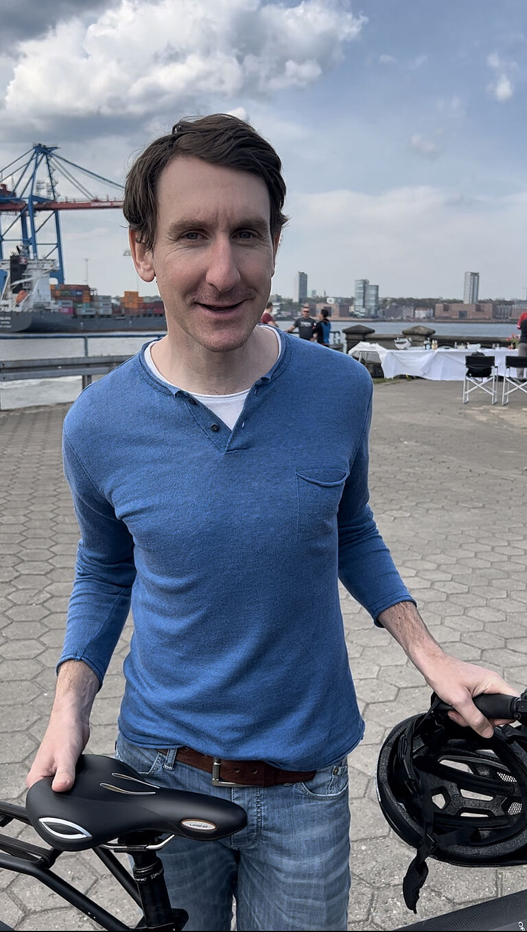 Thomas Springer steht mit seinem Fahrrad am Hafen in Hamburg und lächelt in die Kamera.