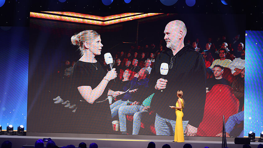 Iris Stelter, links, interviewt in einem Kino in Geesthacht einen Vertreter des Mitgliedsunternehmens BBR Automotive. Im Hintergrund sieht man die Beschäftigten, die zur Live-Stream-Übertragung eingeladen waren. 