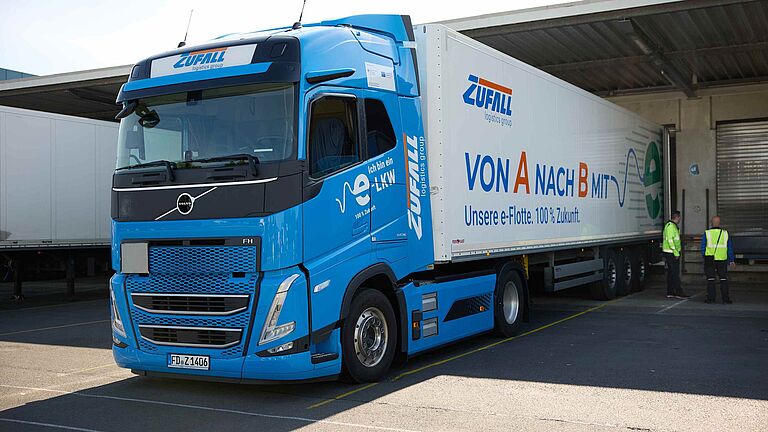 Blauer E-LKW der Zufall Logistics Group mit Aufschrift „Von A nach B mit E“ auf dem Hof.