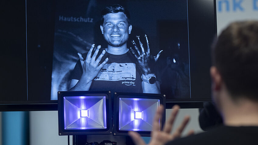 Von hinten ist Niklas Schrage zu sehen, der vor der UV-Kamera und einem Bildschirm sitzt. Er hält seine Hände in die Luft. Auf dem Bildschirm ist die Hand, die er mit Sonnencreme eingecremt hat, in Schwarz gefärbt. 