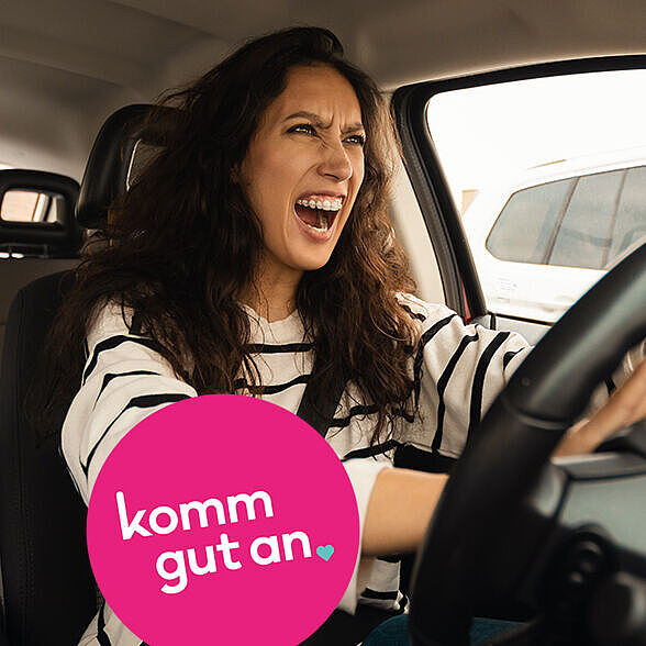Eine junge Frau mit langen dunklen Haaren sitzt am Steuer eines Autos. Sie wirkt wütend, hat den Mund weit geöffnet, als würde sie schreien, und drückt mit beiden Händen auf die Hupe. Im Hintergrund sind andere Fahrzeuge zu sehen. In der linken unteren Bildecke befindet sich ein auffälliger pinker Kreis mit weißer Schrift, auf dem steht: „komm gut an“. Das Bild vermittelt eine angespannte Situation im Straßenverkehr.