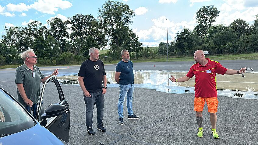Vier Männer stehen am Rande eines Trainingsstrecke für Fahrsicherheit. Rechts erklärt der Fahrtrainer iMarcel n rotem Shirt und leuchtend Organge-farbenen Shorts, worauf es beim Bremsen ankommt.