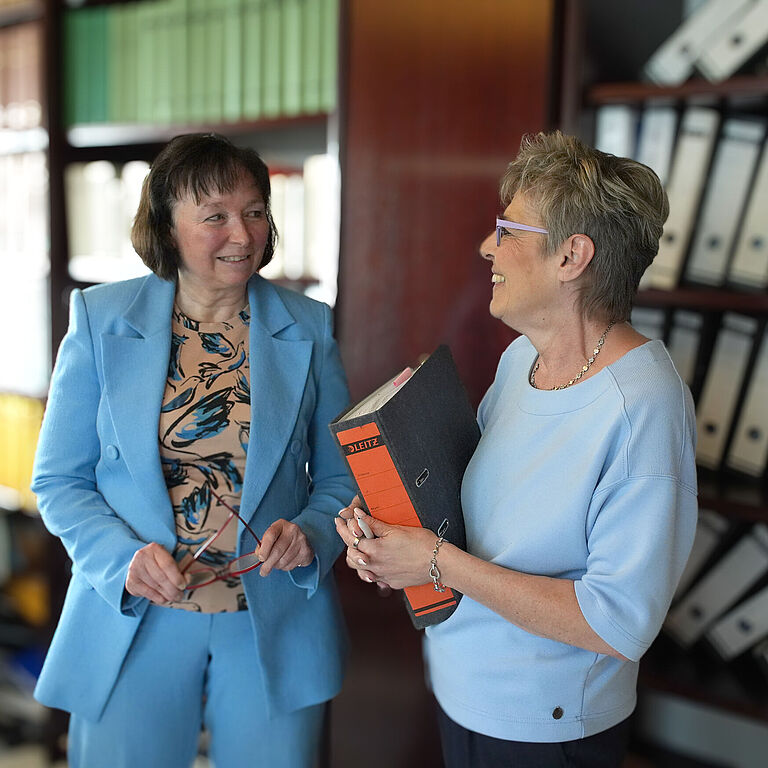 Ute Grötzinger (rechts) und Rechtsanwältin Sylvia Grasser stehen sich vor einem Regal voller Akten gegenüber und sprechen miteinander. Sie lachen sich an.