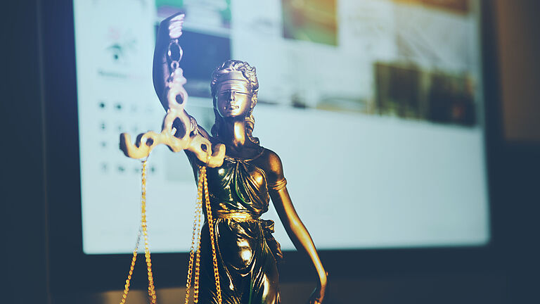 Eine goldene Justizia-Statue ist vor einem Bildschirm zu sehen.