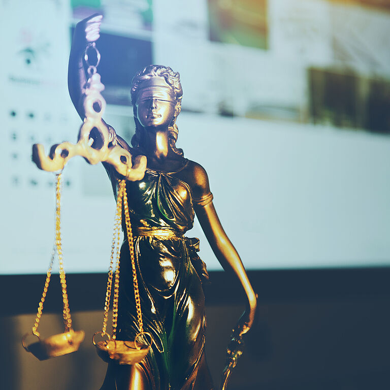 Eine goldene Justizia-Statue ist vor einem Bildschirm zu sehen.