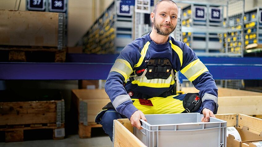 Das Bild zeigt Mitarbeiter Pascal Rudel. Er trägt ein Exoskelett und hebt eine schwere Kiste.