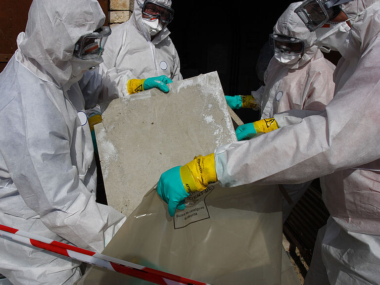 Sanierungsarbeiten durch Arbeiter in weißen Ganzkörperanzügen als Schutzkleidung, die Asbestplatten in einem Plastiksack entsorgen. Davor ein rot-weißes Absperrband.