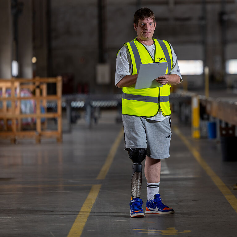 Benjamin Zettl steht in der Nox-Logistikhalle. Er trägt eine kurze Hose. Am rechten Bein ist seine Unterschenkel-Prothese zu sehen. 