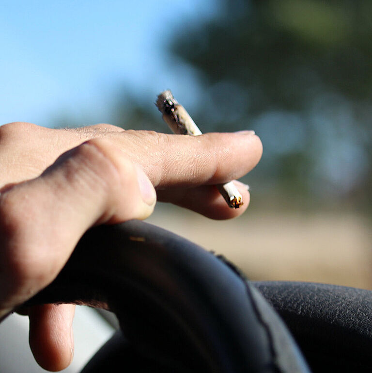Eine Hand liegt auf dem Lenkrad eines Pkw und hält einen Joint in der Hand zwischen zeige- und Mittelfinger. Es ist sonnig, und im Hintergrund ist unscharf ein Baum zu erahnen.