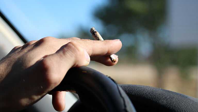 Eine Hand liegt auf dem Lenkrad eines Pkw und hält einen Joint in der Hand zwischen zeige- und Mittelfinger. Es ist sonnig, und im Hintergrund ist unscharf ein Baum zu erahnen. 