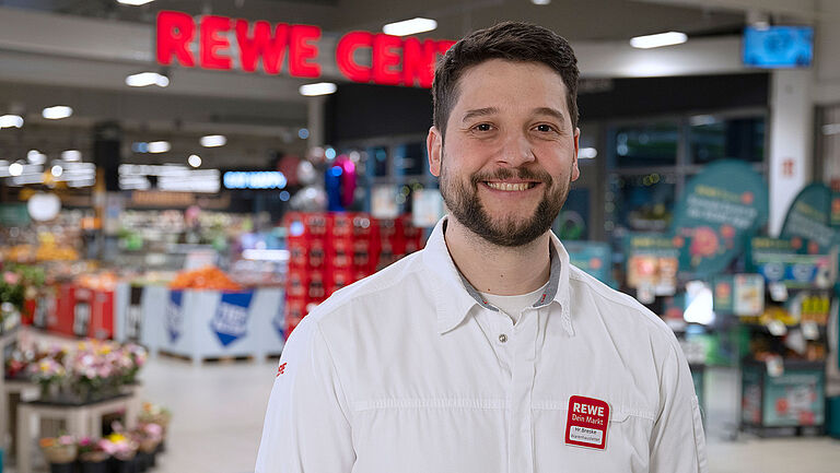 Marktleiter Moritz Breske in weißem Hemd mit Namensschild steht lächelnd in einem gut beleuchteten REWE-Markt vor einem bunten Hintergrund aus Regalen, Obstständen und Schildern. Das Schild über ihm trägt den Schriftzug ‚REWE CENTER‘.