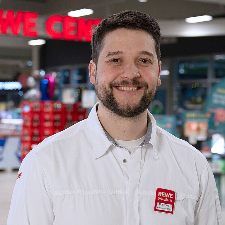 Marktleiter Moritz Breske in weißem Hemd mit Namensschild steht lächelnd in einem gut beleuchteten REWE-Markt vor einem bunten Hintergrund aus Regalen, Obstständen und Schildern. Das Schild über ihm trägt den Schriftzug ‚REWE CENTER‘.