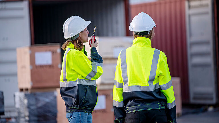 Eine Frau und ein Mann mit Warnjacken und Schutzhelmen stehen mit dem Rücken zur Kamera und schauen auf Container, sie spricht in ein Funksprechgerät.