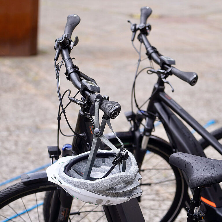 Am Lenker eines schwarzen Pedelecs hängt ein silberner Fahrradhelm. Nur der Lenker des Rades und zwei daneben stehender Fahrräder sind zu sehen.