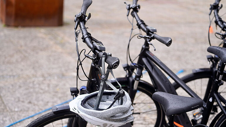 Am Lenker eines schwarzen Pedelecs hängt ein silberner Fahrradhelm. Nur der Lenker des Rades und zwei daneben stehender Fahrräder sind zu sehen.