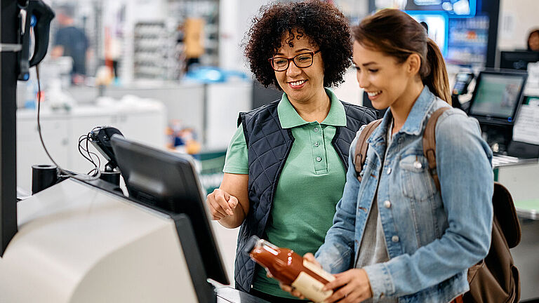 Zwei Frauen stehen an einer Self-Checkout-Kasse im Supermarkt; eine Mitarbeiterin erklärt einer Kundin den Scanvorgang eines Produkts.