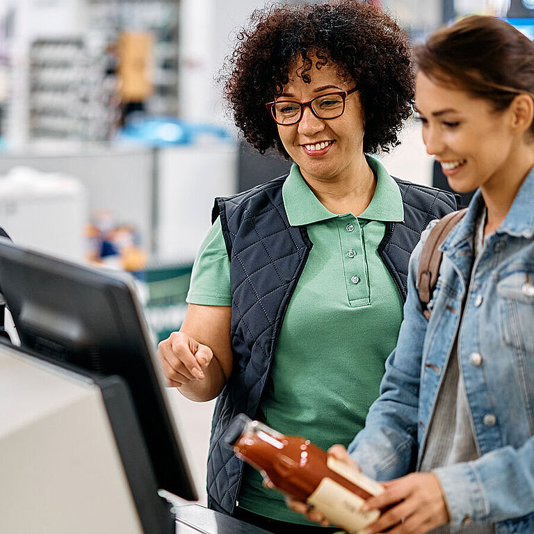 Zwei Frauen stehen an einer Self-Checkout-Kasse im Supermarkt; eine Mitarbeiterin erklärt einer Kundin den Scanvorgang eines Produkts.