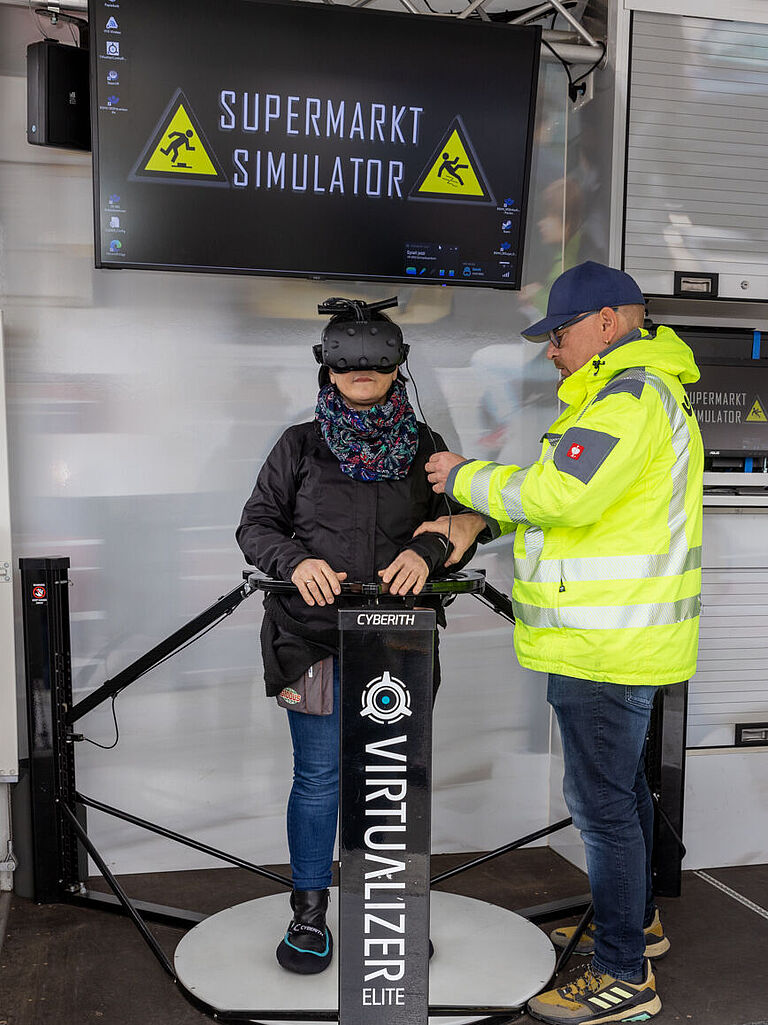 Betriebsratsvorsitzende Tanja Meyer steht im BGHW mobil in einem Stahlgestell, ein sogenannter Virtualizer. Sie hat eine VR-Brille an, die sie in die virtuelle Welt eines Supermarkt-Lagers eintauchen lässt. Rechts von ihr steht ein Mann in gelb-leuchtender Jacke. Der Die BGHW-Trainer erklärt ihr, wie die Technik funktioniert. 