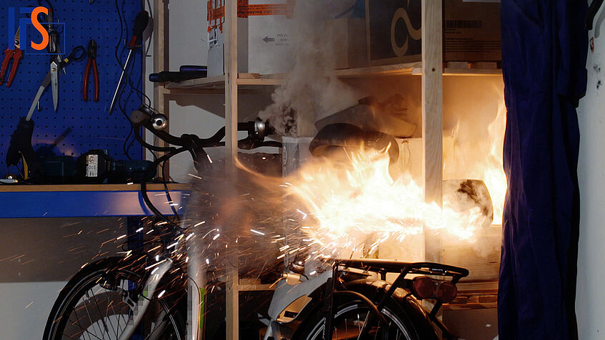 In einem Kellerraum lehnt ein E-Bike an einem Regal und im Regal hat der Akku Feuer gefangen.