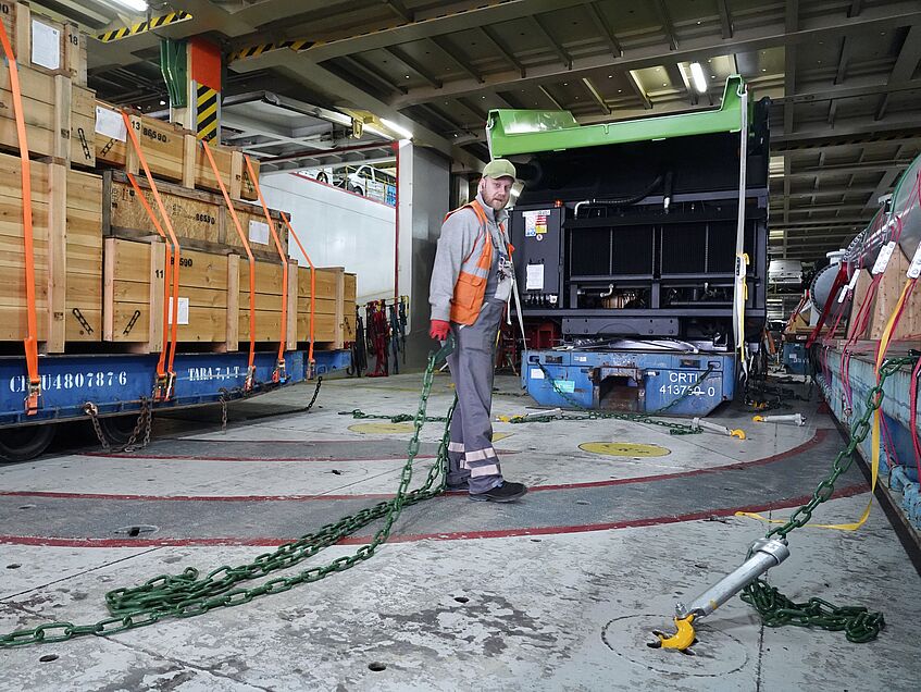 Hafenarbeiter mit Sicherungsketten an Deck des Frachtschiffs. Rechts und links von ihm sieht man schwere Ladung.