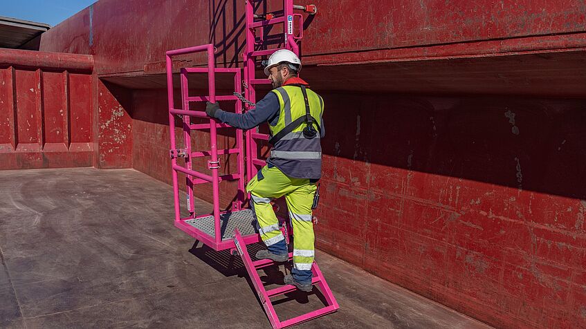 Mann in Sicherheitskleidung betritt über eine Leiterkonstruktion den Ladebereich eines Transportschiffes.