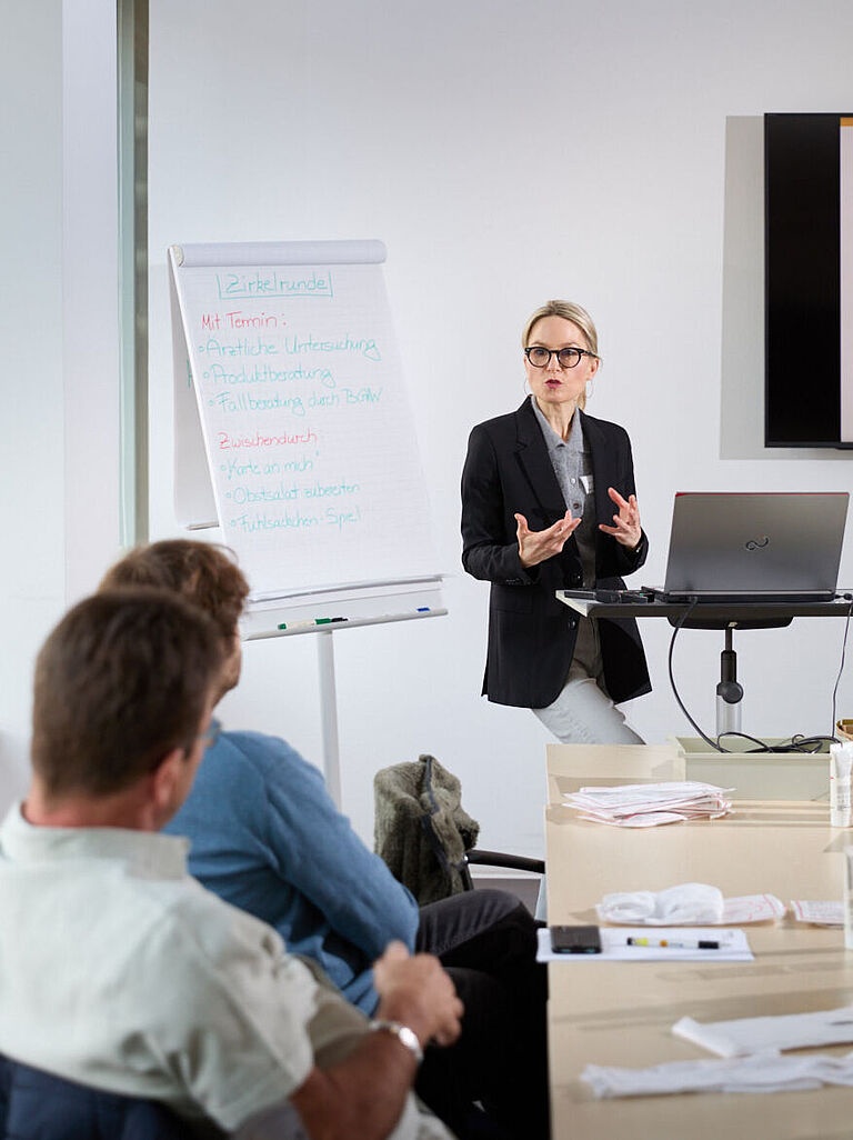 Die Hautärztin steht zwischen Laptop und Flipchart und hält einen Vortrag. Zwei Seminarteilnehmende hören ihr zu. 