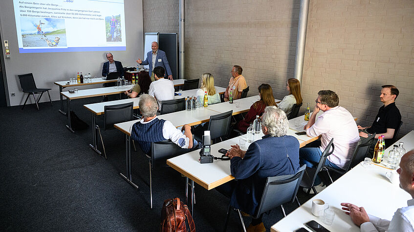 Eine Workshop-Situation während des Reha-Forums: Jeweils zwei Tische hintereinander sitzen die Teilnehmenden eines Workshops, die mit dem Rücken zur Kamera sitzen. Rechts zum Ende der Tischreihen ist eine beige Wand. Von den beiden Referenten sitzt eine Person, die andere steht und gestikuliert. Im Hintergrund an der Wand ist eine Power-Point-Präsentation zu sehen. Tischreihen 