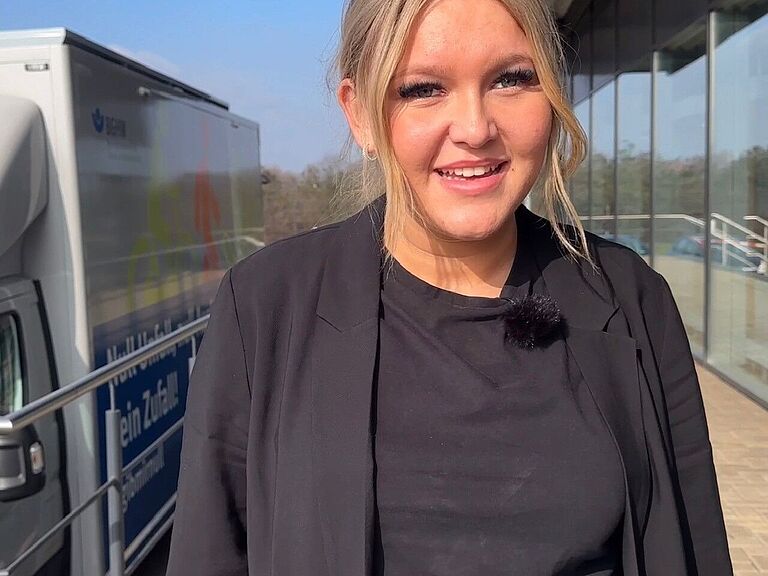 Eine junge Frau mit blonden Haaren schaut in die Kamera. Hinter ihr sind Ausschnitte eines Fahrzeugs und Gebäudes zu sehen. 