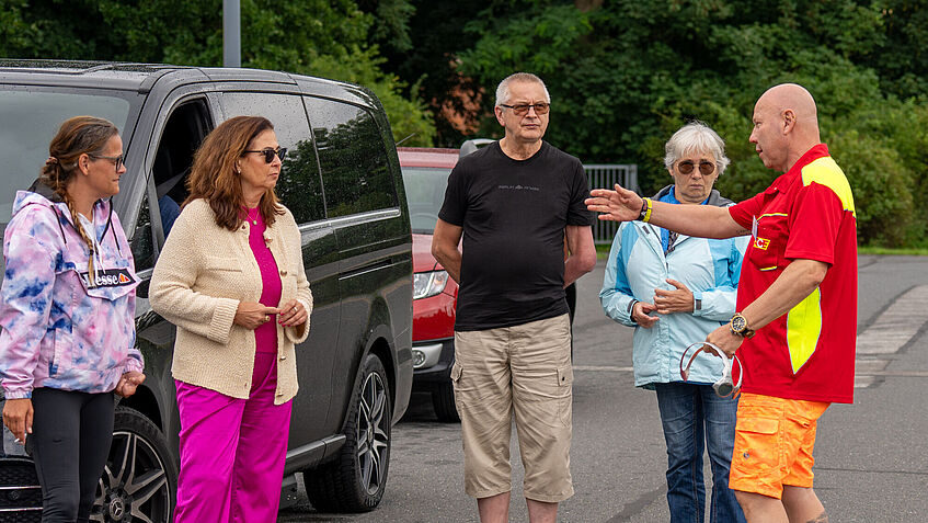 Fünf Person stehen neben einem Fahrzeug, drei Frauen und zwei Männer. Rechts steht gestikulierend Fahrtainer Marcel, in rotem Shirt und in leuchtend Orange-farbener Hose. 