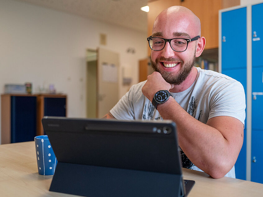 Der BGHW-Versicherte sitzt an einem Tischu und schaut auf sein vor ihm aufgestelltes Tablet. Er greift sich lachend mit der linken Hand in den Bart. 
