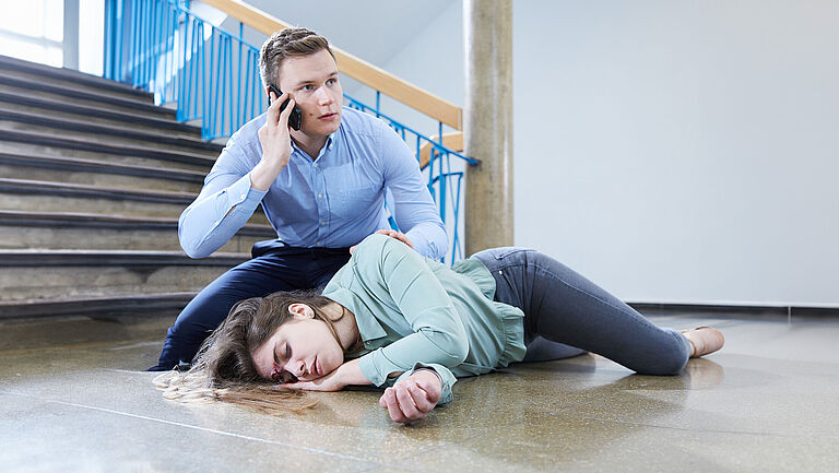 Ein Mann kniet am Fußende einer Treppe bei einer bewusstlosen Frau und telefoniert mit besorgtem Gesichtsausdruck