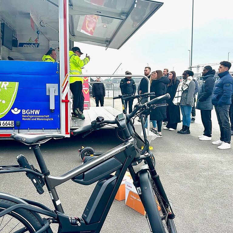 Ein Mann steht mit einem Pedelec vor einer Gruppe von Menschen. Links von der Gruppe ist das BGHW-"mobil", ein Anhänger mit Display, Helmen und einem Laufband.