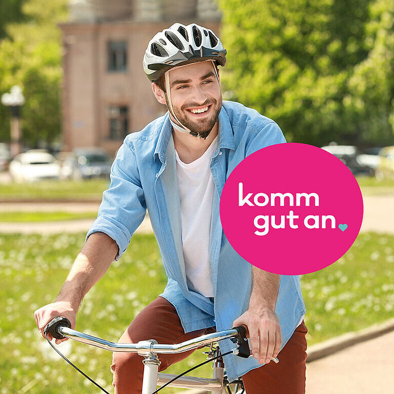Ein lächelnder Radfahrer mit Fahrradhelm fährt durch eine Parkanlage. Im Vordergrund befindet sich ein pinker Kreis mit dem weißen Schriftzug „komm gut an“. Im Hintergrund sind Bäume, parkende Autos und ein Gebäude unscharf zu sehen.