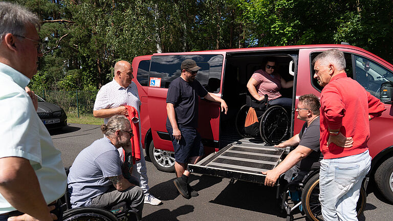 Die Teilnehmenden stehen um einen roten Kleinbus, an dessen rechter Seitentür eine Rampe hochgefahren wurde. Darauf steht Rollstuhlfahrerin Kristin. Gemeinsam tauschen sie sich über die Fahrzeug-Umbauten aus. ber ihre Pkw-Umbauten aus und werden individuell beraten.