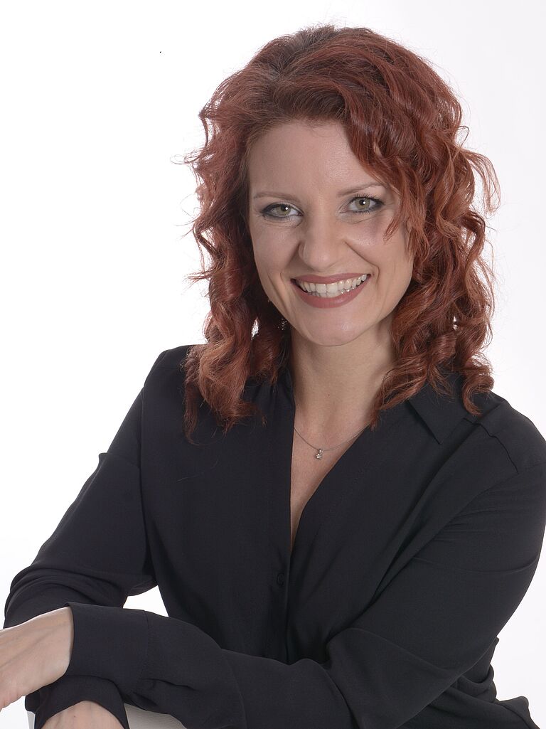 Katja Sessig, BGHW-Referentin Berufskrankheiten und digitale Rehabilitation, schaut freundlich in die Kamera. Sie hat die Arme verschränkt, trägt eine schwarze Bluse und hat rotes langes Haar.