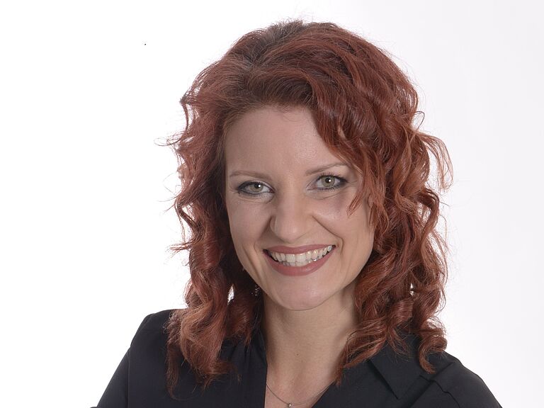 Katja Sessig, BGHW-Referentin Berufskrankheiten und digitale Rehabilitation, schaut freundlich in die Kamera. Sie hat die Arme verschränkt, trägt eine schwarze Bluse und hat rotes langes Haar.