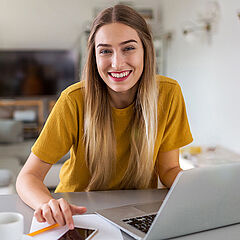 Eine junge Frau sitzt lächelnd an einem Laptop, der auf einem Tisch in ihrem Wohnzimmer steht.