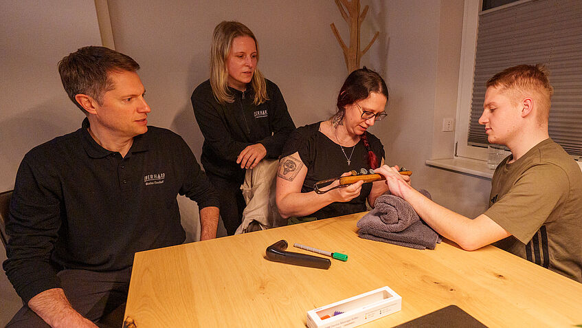 Lars (rechts) sitzt am heimischen Esstisch. Ihm gegenüber sitzt Ergotherapeutin Iris Stenzel und hält einen Stab unter seine linke Hand, die er ihr entgegenstreckt. Links von ihr sind Lars' Eltern, die der Behandlung gespannt zuschauen. 