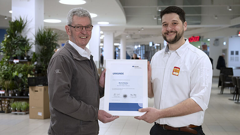 Frank Kiowski und Moritz Breske stehen in einem gut beleuchteten Einkaufszentrum, zu dem ein REWE Center gehört. Moritz Breske auf der rechten Seite trägt ein weißes Hemd mit einem REWE-Namensschild und hält gemeinsam mit BGHW-Aufsichtsperson Frank Kiowski eine Urkunde. Beide lächeln in die Kamera, während im Hintergrund Pflanzen und Geschäftsräume zu sehen sind.