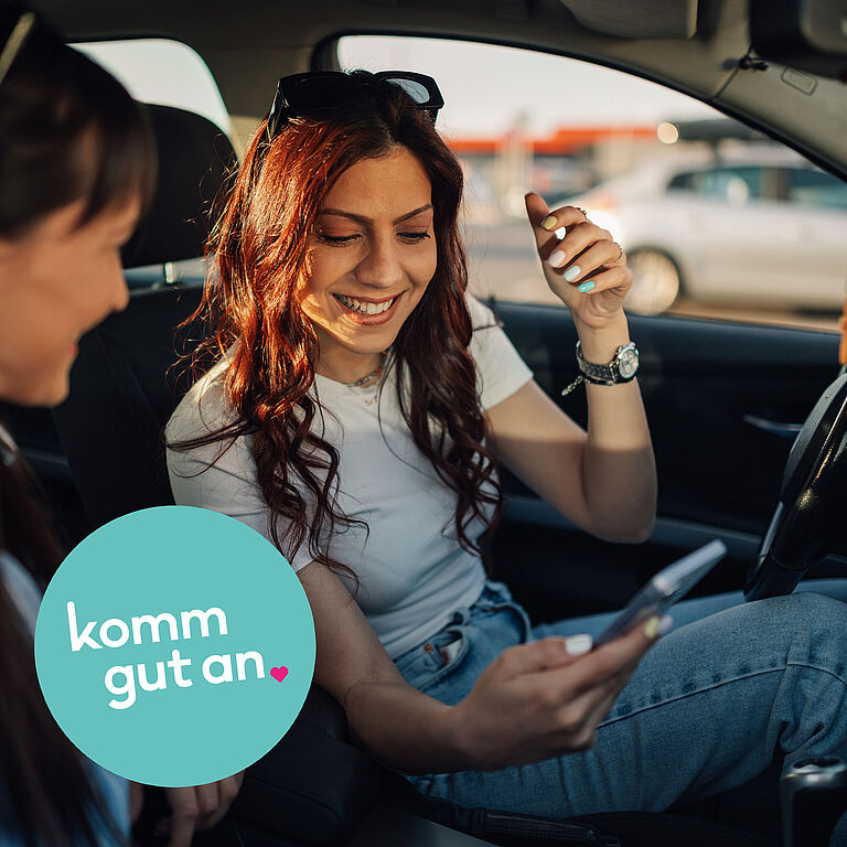 Zwei Personen sitzen in einem geparkten Auto. Die Person auf dem Fahrersitz hält ein Smartphone in der Hand und unterhält sich mit der Beifahrerin. Auf der Bildfläche ist zudem ein rundes Logo mit dem Schriftzug ‚komm gut an‘ zu sehen.