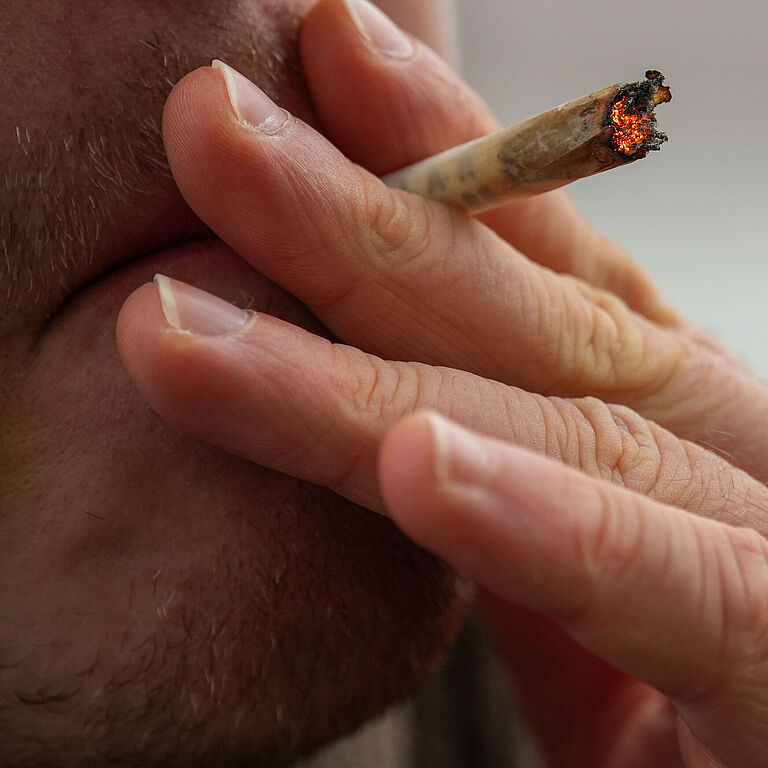  Eine Person raucht einen Joint, der zwischen Zeige- und Mittelfinger steckt. Man sieht nur einen Ausschnitt des Gesichtes, das durch die linke Hand der Person verdeckt ist, die an dem glühenden Joint zieht.