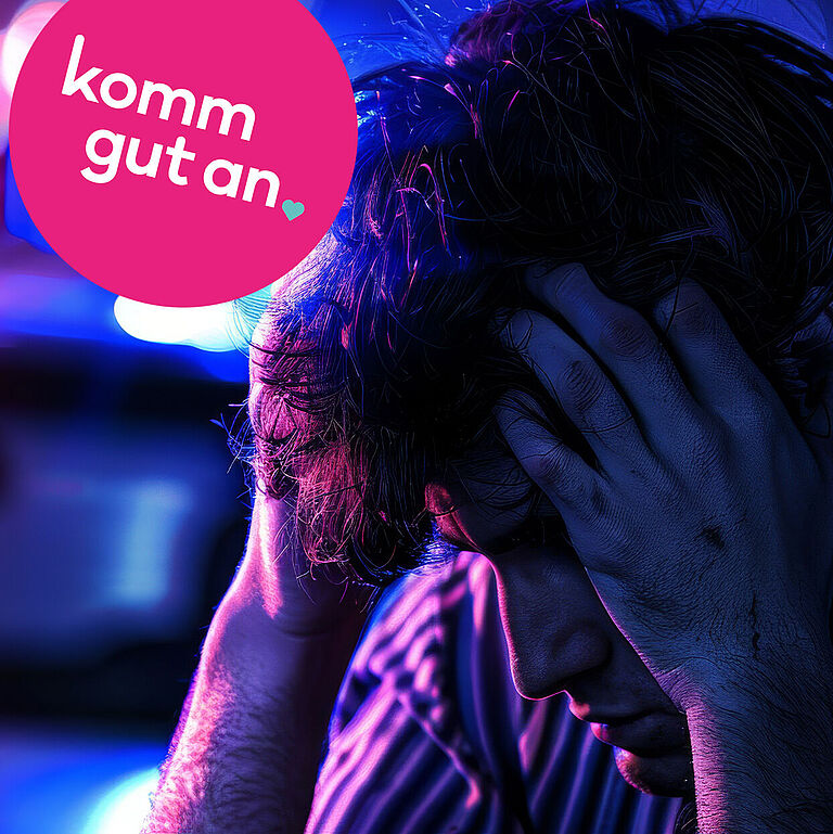 Verzweifelter junger Mann sitzt nachts am Straßenrand vor einem Polizeiwagen mit blau-roten Einsatzlichtern und hält den Kopf in den Händen. Im Bild befindet sich ein pinker Kreis mit dem Slogan ‚komm gut an‘. Symbolfoto für Unfall, Notfall, Stress im Straßenverkehr und Verkehrssicherheit.