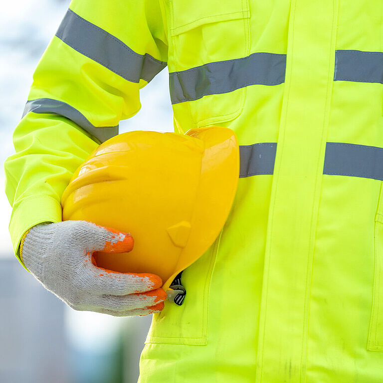 Zu sehen ist der Oberkörper einer Person, die eine neongelbe Warnjacke trägt, Sicherheitshandschuhe und einen Schutzhelm unter dem rechten Arm festhält.