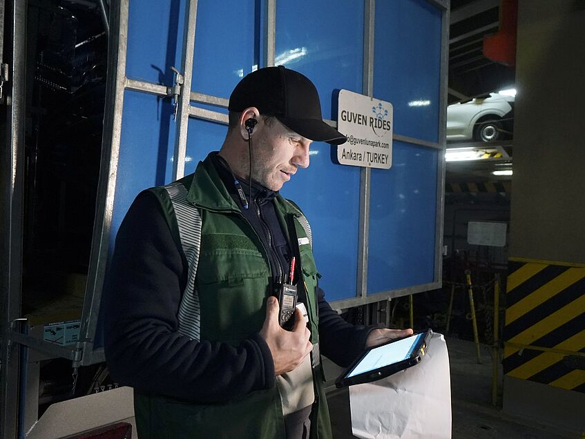 Hafenarbeiter mit Funkgerät und Tablet auf Fahrzeugdeck eines Frachtschiffs.