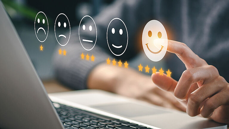 Abstimmungssituation. Imaginäre Smileys von schlecht (1 Stern) bis sehr gut (5 Sterne) werden angezeigt. Hände liegen an einem Laptop. Der linke Zeigefinger drückt auf den 5 Sterne Smiley für sehr gut.