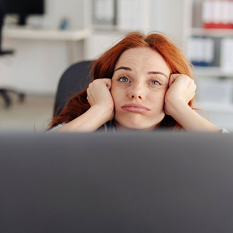 Junge Frau mit roten Haaren stützt ihren Kopf mit beiden Händen, blickt erschöpft und gelangweilt über einen Laptop hinweg, im Hintergrund ein unscharfes Büro mit Regalen und Aktenordnern.