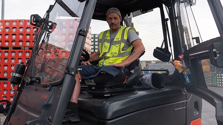 Ein Mann in gelber Warnweste sitzt auf einem Gabelstapler und schaut in die Kamera. Ihm fehlt sein linkes Bein. Recht hinter dem Sitz liegt seine Prothese.