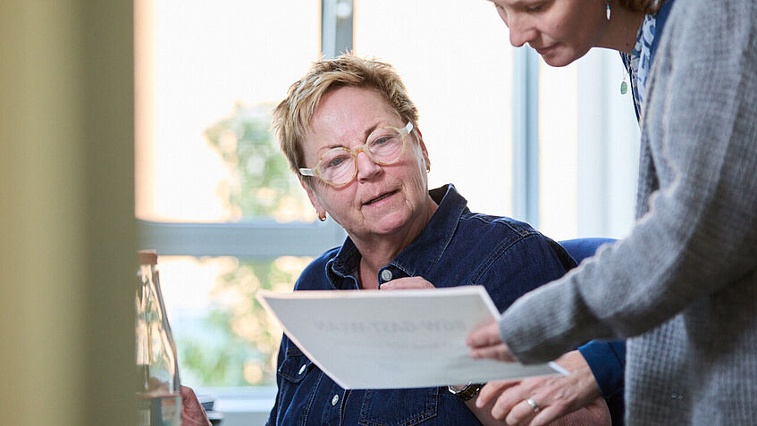 Die am Schreitisch sitzende Seminarleiterin schaut auf ein Papier, das ihr die Gesundheitspädagogin zeigt, die neben ihr steht. 