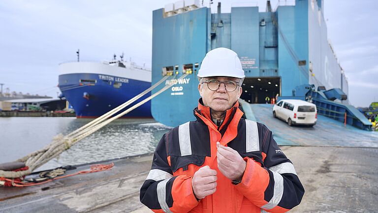 Aufsichtsperson der BGHW mit Helm und Warnjacke vor Autotransportschiff im Hafen. In der Hand hält er eine Otoplastik.